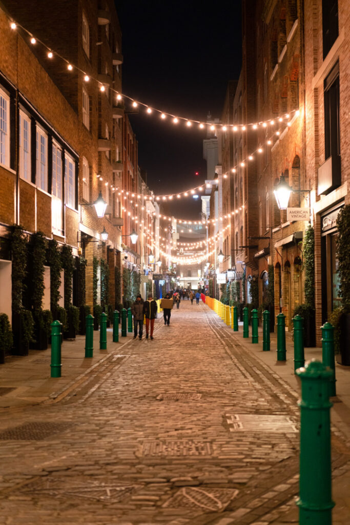 A quiet cobbled street in London at night, illuminated by decorative string lights and lined with historic buildings.
