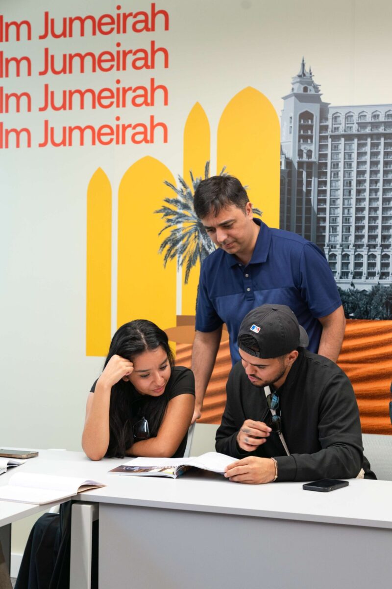 Teacher assisting two students during an English lesson at EC Dubai, with classroom materials and Dubai-inspired wall artwork in the background.