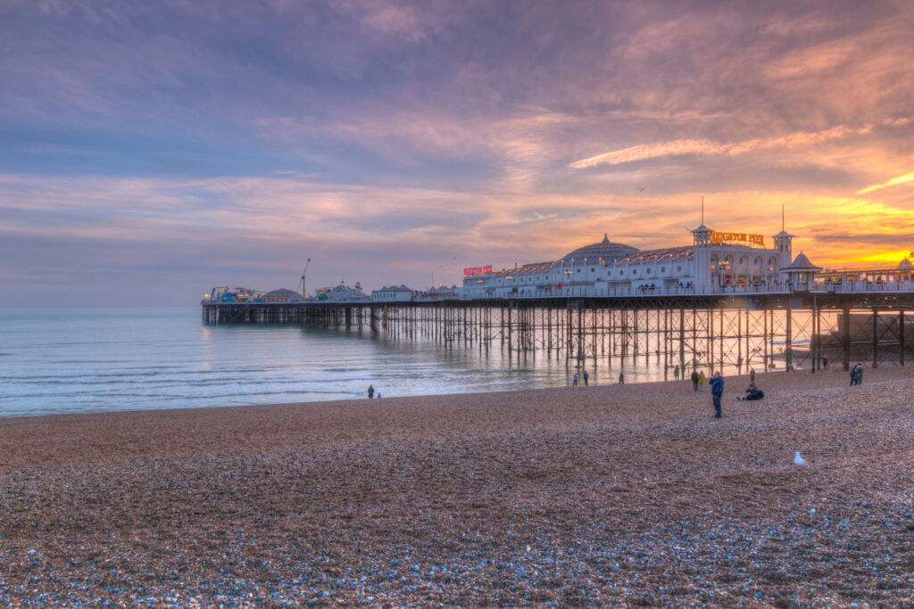 Il celebre Molo di Brighton al tramonto, con la spiaggia di ciottoli, il mare calmo e un cielo dai colori caldi.